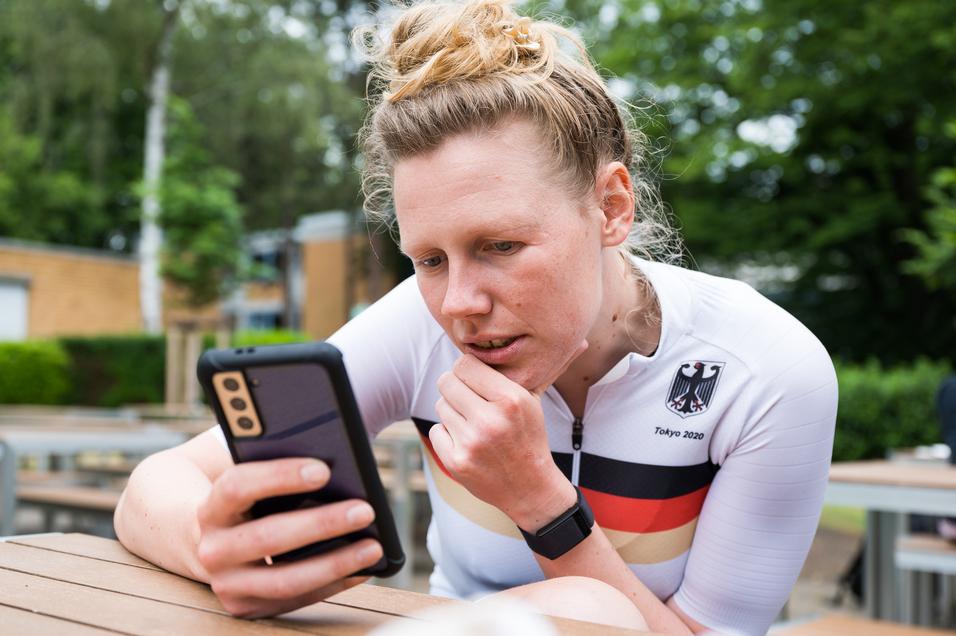 Athletin im weißen Trikot mit schwarzem und rotem Streifen schaut konzentriert auf ihr Smartphone in einer Außenanlage.